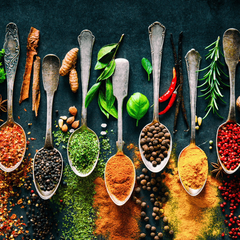 An overhead flat-lay arrangement of vintage silver spoons filled with whole and ground spices on a dark textured background. Visible spices include red chili peppers, green basil leaves, coriander seeds, cinnamon sticks, vanilla pods, rosemary, and powders of turmeric, paprika, cumin, and chili.