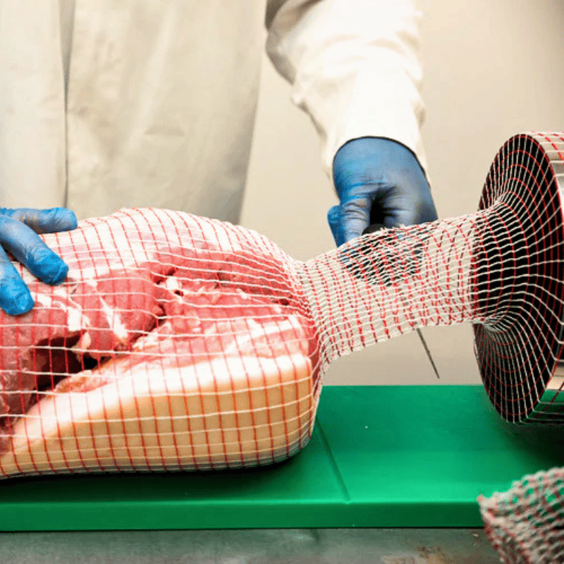 A person wearing white protective clothing and blue gloves handles a large piece of raw meat wrapped in red-and-white mesh netting on a green surface.