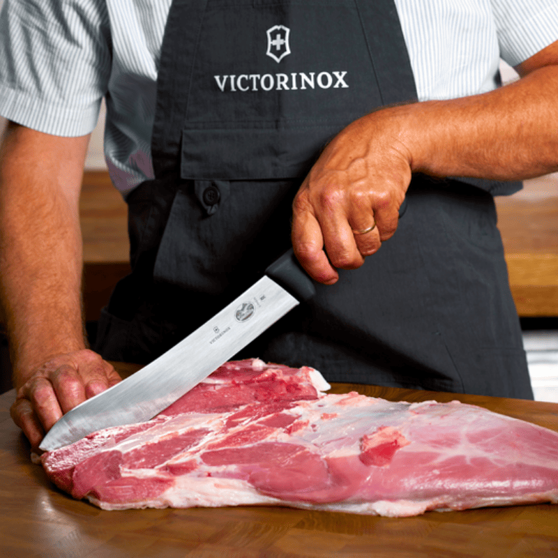 A person wearing a black Victorinox apron uses a large chef's knife to cut a large piece of raw meat on a wooden table.